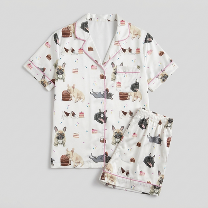 Women's satin pajama set with cream, fawn and gray French bulldogs between birthday cakes and strawberries on white - flat lay