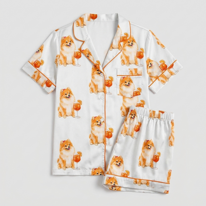 Women's satin pajama set with Pomeranians beside orange spritz cocktails on white with orange piping - flat lay