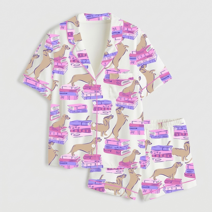 Women's satin pajama set with tan dachshunds sprawled on stacks of pink and purple books on white - flat lay