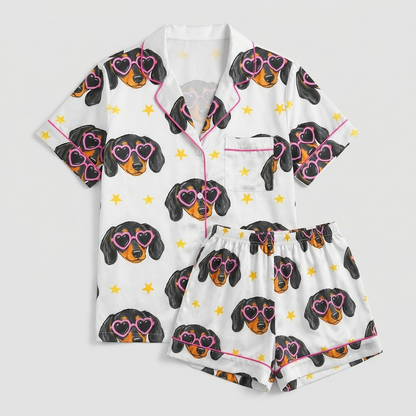 Women's satin pajama set with black-and-tan dachshund faces in pink heart sunglasses and gold stars on white - flat lay