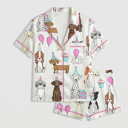 Women's satin pajama set with dogs in party hats beside pink balloons, cupcakes and wrapped gifts on white - flat lay