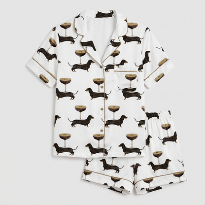 Women's long-sleeve satin pajama set with black dachshunds beside gold-stemmed coupe glasses on white - flat lay